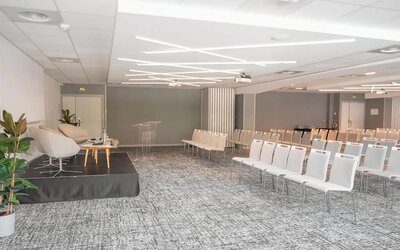 1. Large conference room with rows of white chairs, a presentation stage, podium, plants, and modern lighting at Holiday Inn Toulouse Airport.