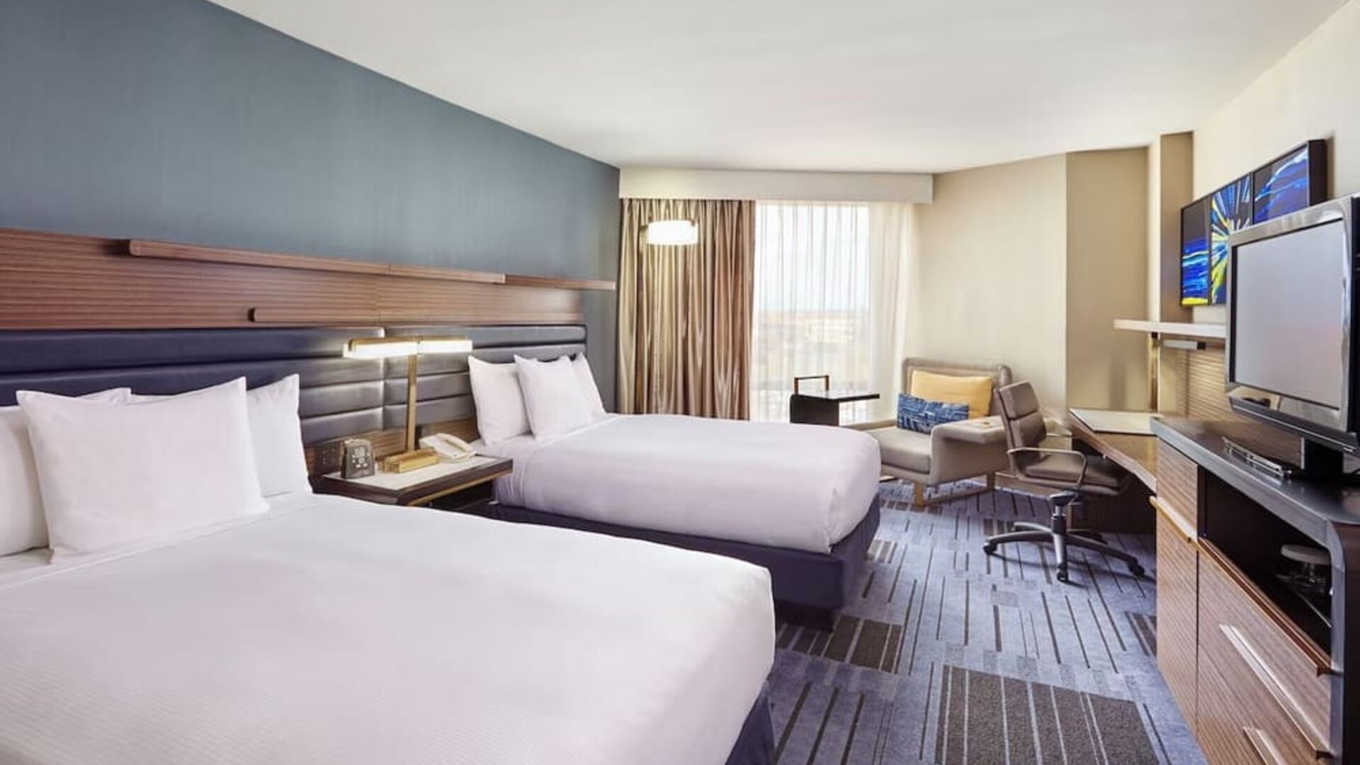 1. Guest room with two double beds, blue accent wall, bedside table, work desk, lounge chair, television, and large windows at Hilton Newark Airport.