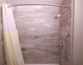 Bathtub and shower with wood-look tiled walls, curved shower rod, showerhead, grab bar, and white shower curtain at At Home Inn & Suites.