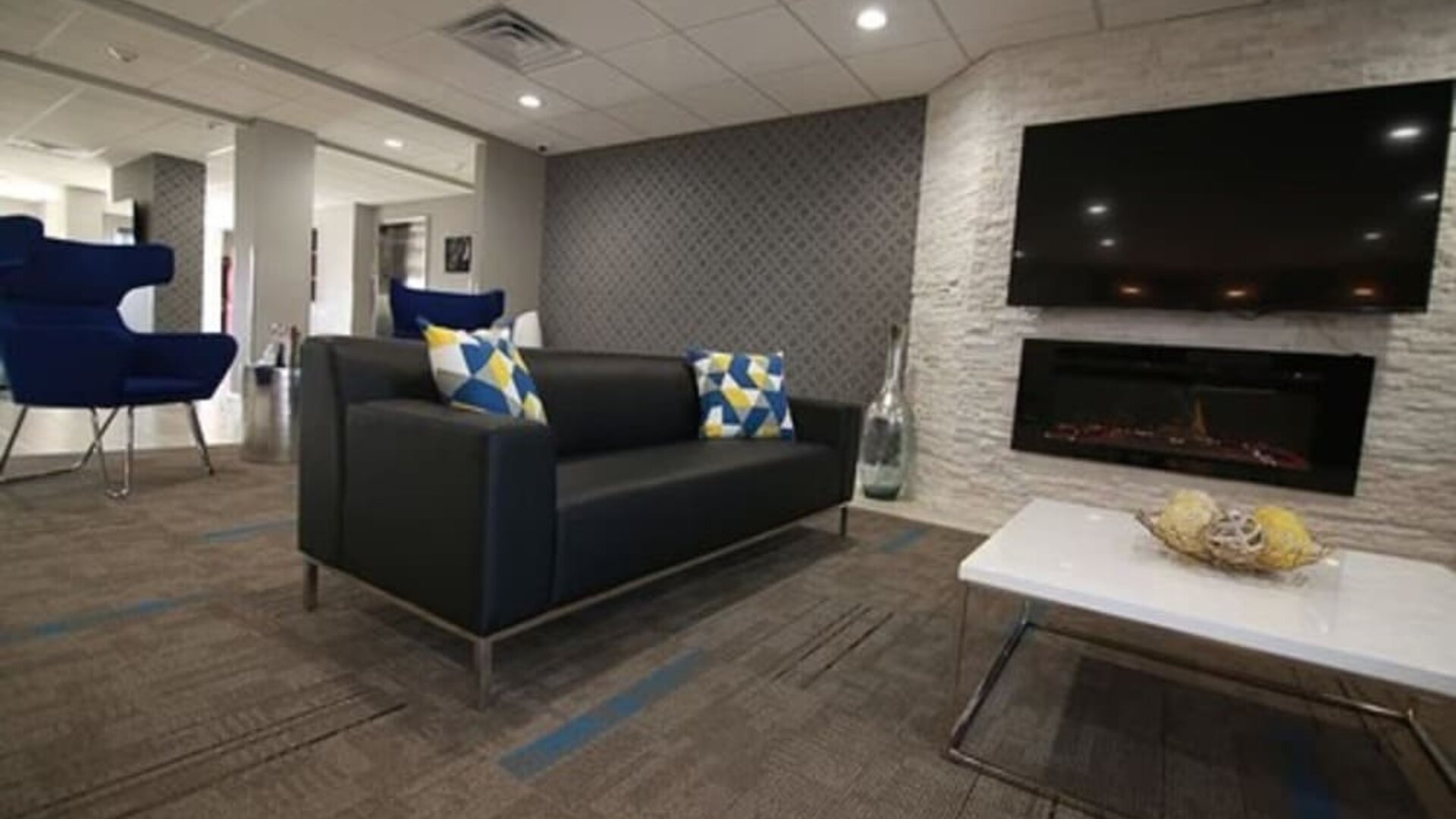 Hotel lounge with black sofas, blue armchairs, a fireplace, wall-mounted television, and white coffee tables at At Home Inn & Suites.
