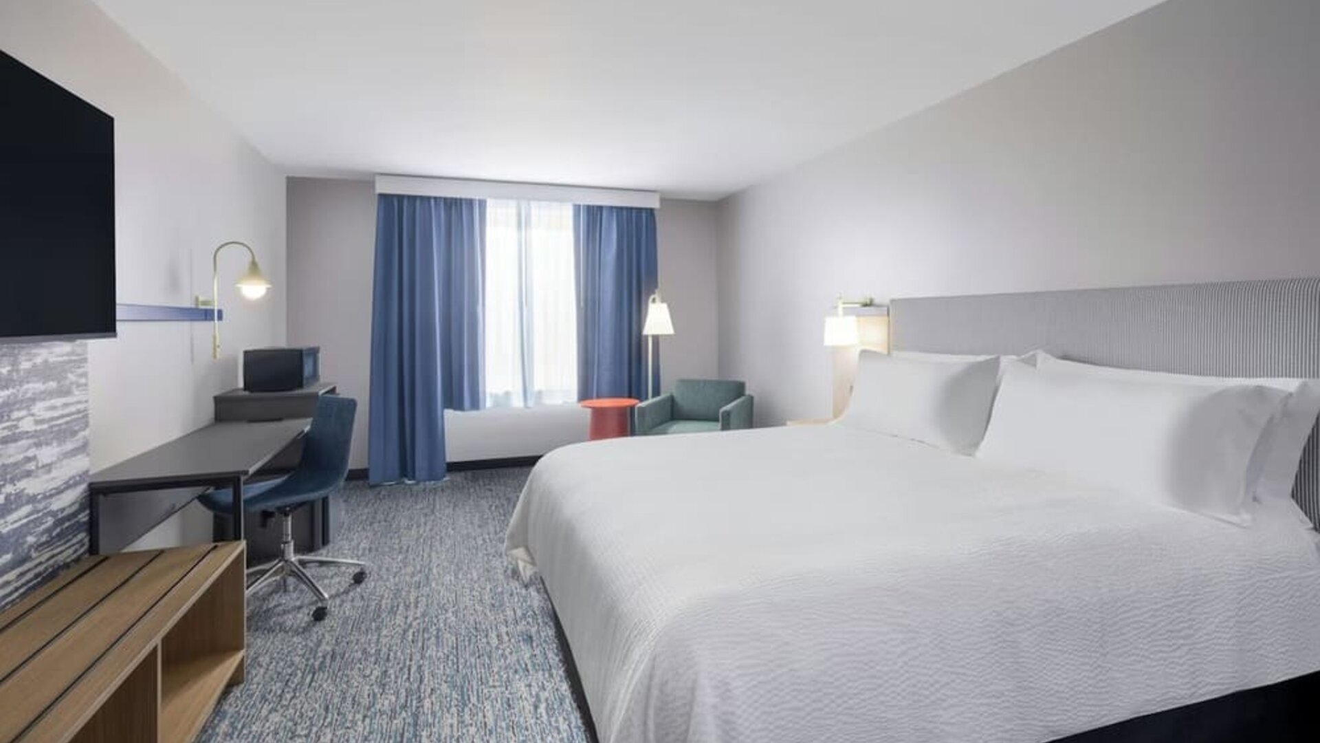 1. Spacious guest room with a king bed, white linens, desk and chair, television, seating area, blue curtains, and patterned carpet at Garner by IHG.