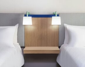 Two hotel beds with white linens, upholstered headboards, wood nightstand, and wall-mounted reading lamps at Garner by IHG.