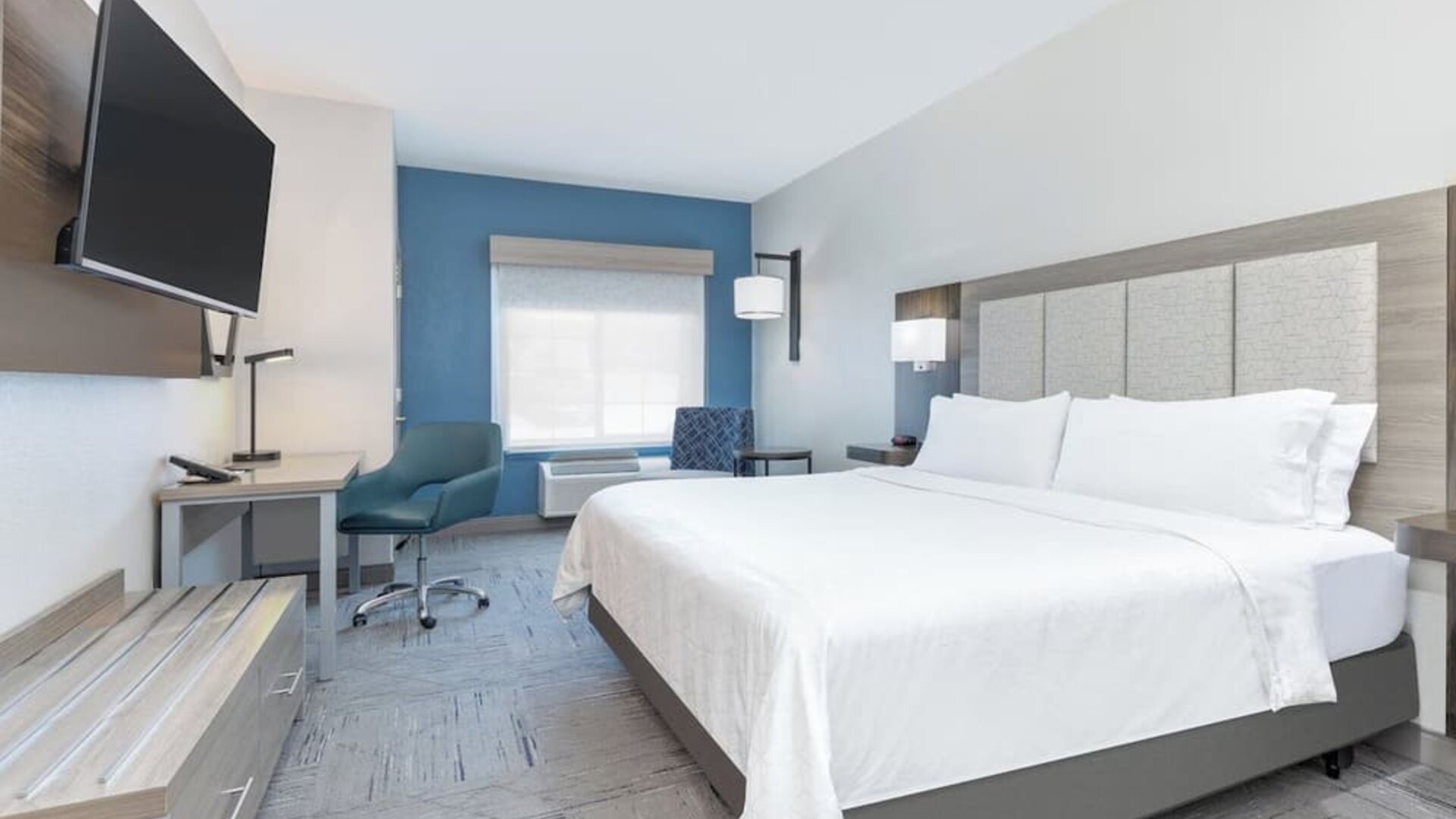 King guest room with a large bed, white bedding, blue accent wall, desk, armchair, and wall-mounted TV at Holiday Inn Express & Suites Granbury.