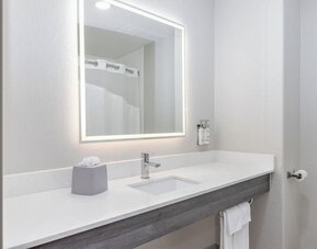 Bathroom vanity with a large illuminated mirror, white countertop, integrated sink, and wall-mounted toiletries at Holiday Inn Express & Suites Granbury.