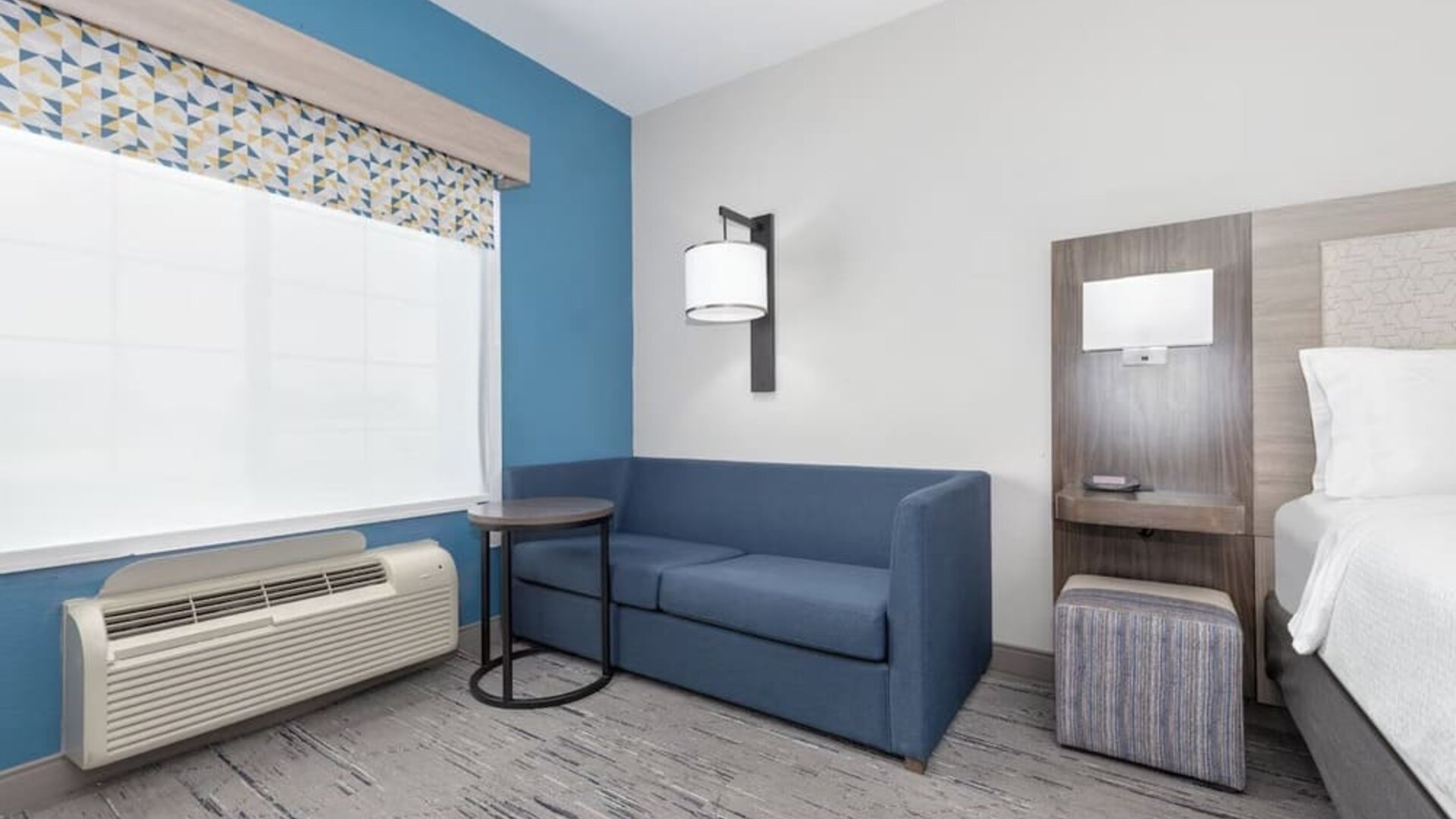 Guest room seating area with a blue sofa, side table, patterned window valance, and bedside area at Holiday Inn Express & Suites Granbury.