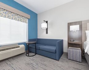 Guest room seating area with a blue sofa, side table, patterned window valance, and bedside area at Holiday Inn Express & Suites Granbury.
