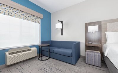 Guest room seating area with a blue sofa, side table, patterned window valance, and bedside area at Holiday Inn Express & Suites Granbury.