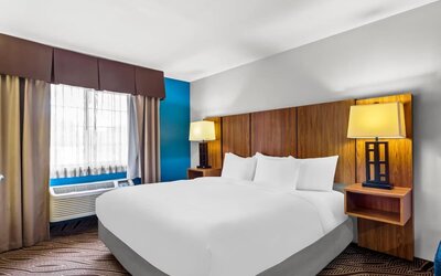 Comfortable king guest room featuring a large bed with white linens, wooden headboard, bedside lighting, blue accent wall, window, air-conditioning unit, and contemporary décor.