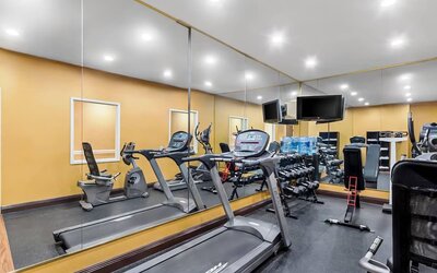 Compact hotel fitness center with treadmills, stationary bike, dumbbell rack, strength-training equipment, wall-mounted televisions, and large mirrored walls.