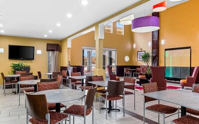 Bright hotel dining and lounge area with multiple tables and chairs, colorful pendant lights, wall-mounted television, decorative plants, and contemporary seating beside a modern water feature.