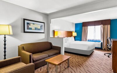Spacious hotel guest room with a king bed, separate sitting area with brown sofa and armchair, coffee table, work desk, large window, and bright blue accent wall.