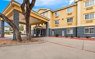 Exterior view of a multi-story hotel with beige and gray façade, covered entrance and driveway, mature trees, and rows of guest room windows.