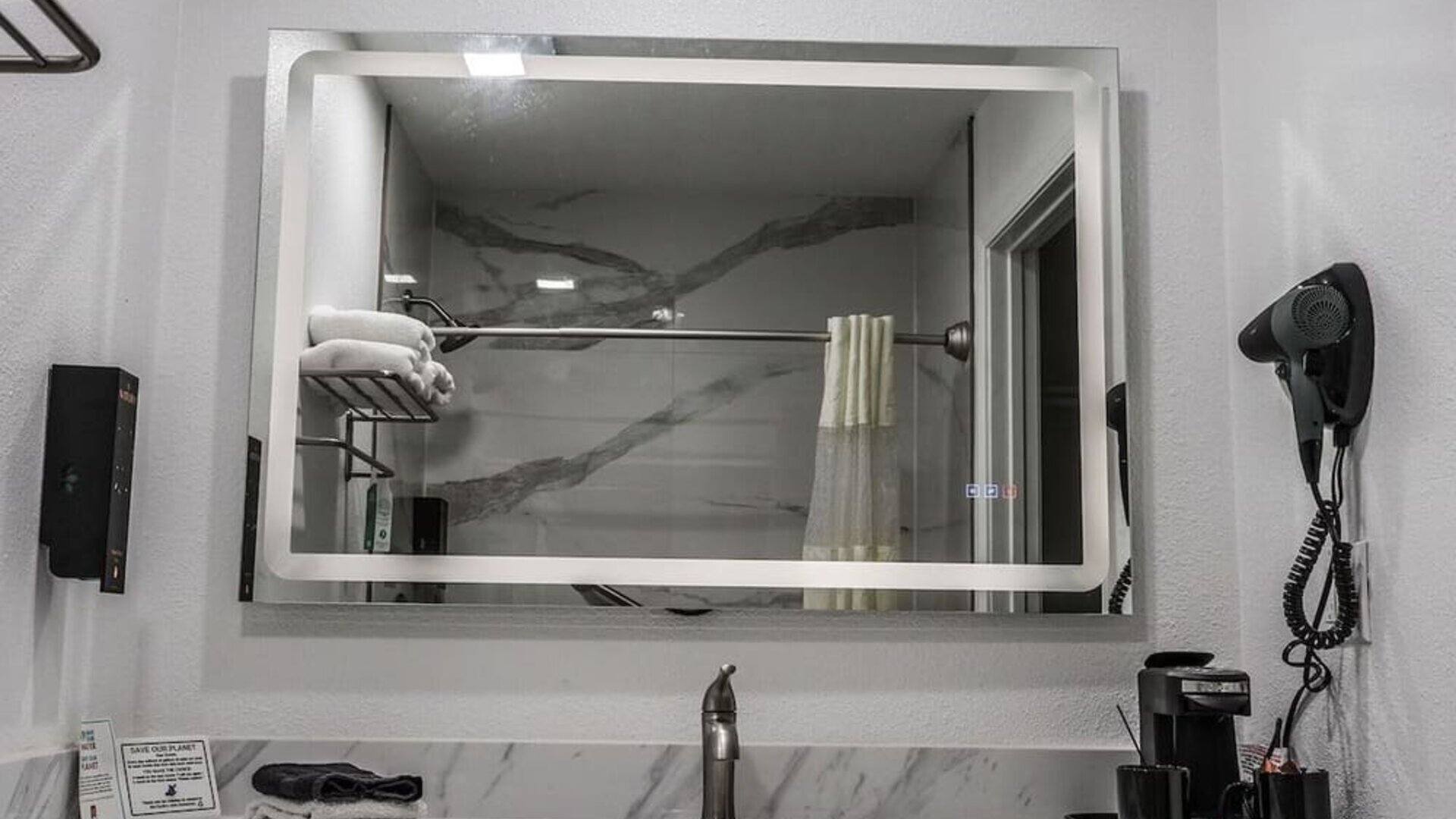 Bathroom vanity with a large illuminated mirror, marble-look backsplash, sink, hairdryer, towels, and in-room coffee setup at Western Inn.