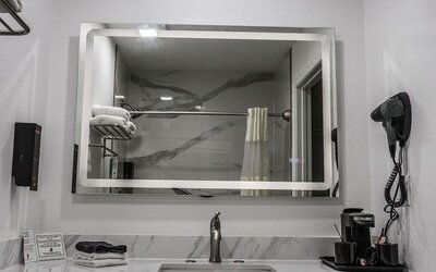 Bathroom vanity with a large illuminated mirror, marble-look backsplash, sink, hairdryer, towels, and in-room coffee setup at Western Inn.