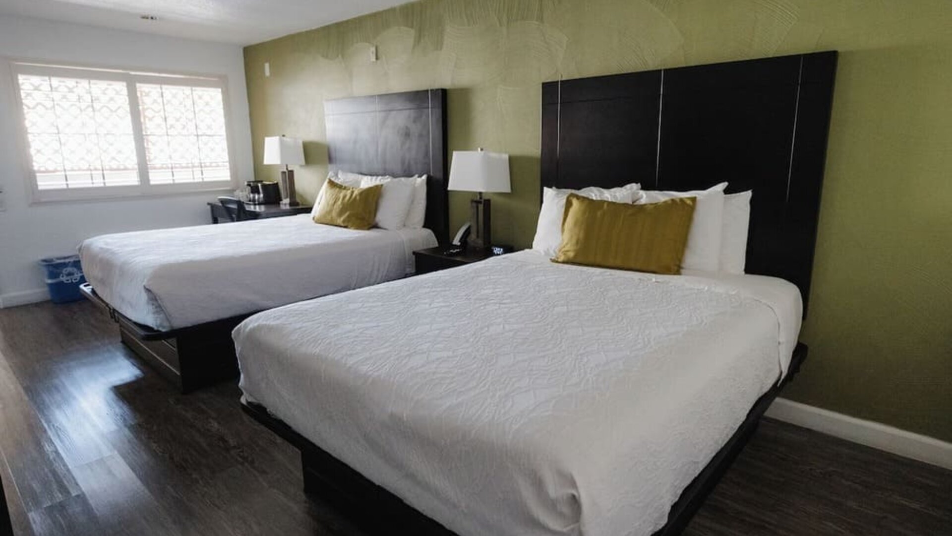 Guest room with two beds, white bedding, green accent wall, dark wood flooring, bedside lamps, and large windows at Western Inn.