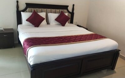 Spacious double bed featuring crisp white bedding, burgundy accent pillows, a quilted runner, an upholstered wooden headboard, and bedside furniture.