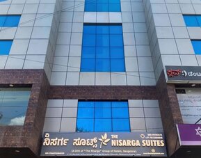 Multistory exterior of The Nisarga Suites in Bangalore featuring reflective blue glass windows, silver facade panels, brown stone accents, and hotel signage.
