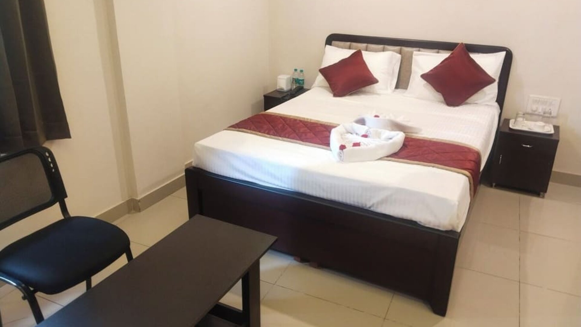Comfortable double bed guest room featuring white linens, burgundy decorative pillows, matching bed runner, folded towels, bedside cabinets, and a seating area.