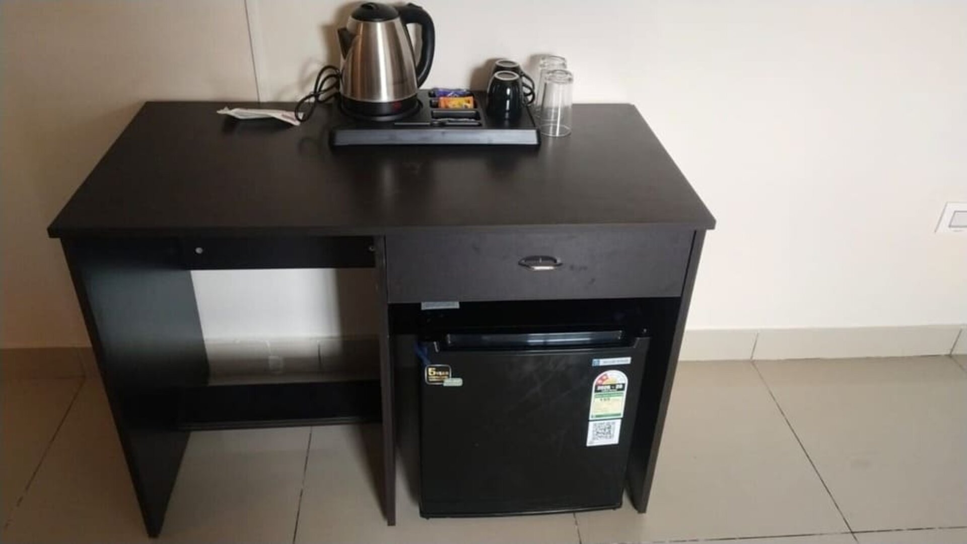 Black wooden refreshment station at The Nisarga Suites featuring an electric kettle, mugs, drinking glasses, beverage supplies, and a mini refrigerator.