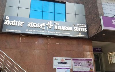 Exterior entrance of The Nisarga Suites in Bangalore displaying the hotel's illuminated English and Kannada signage beneath reflective blue glass windows.