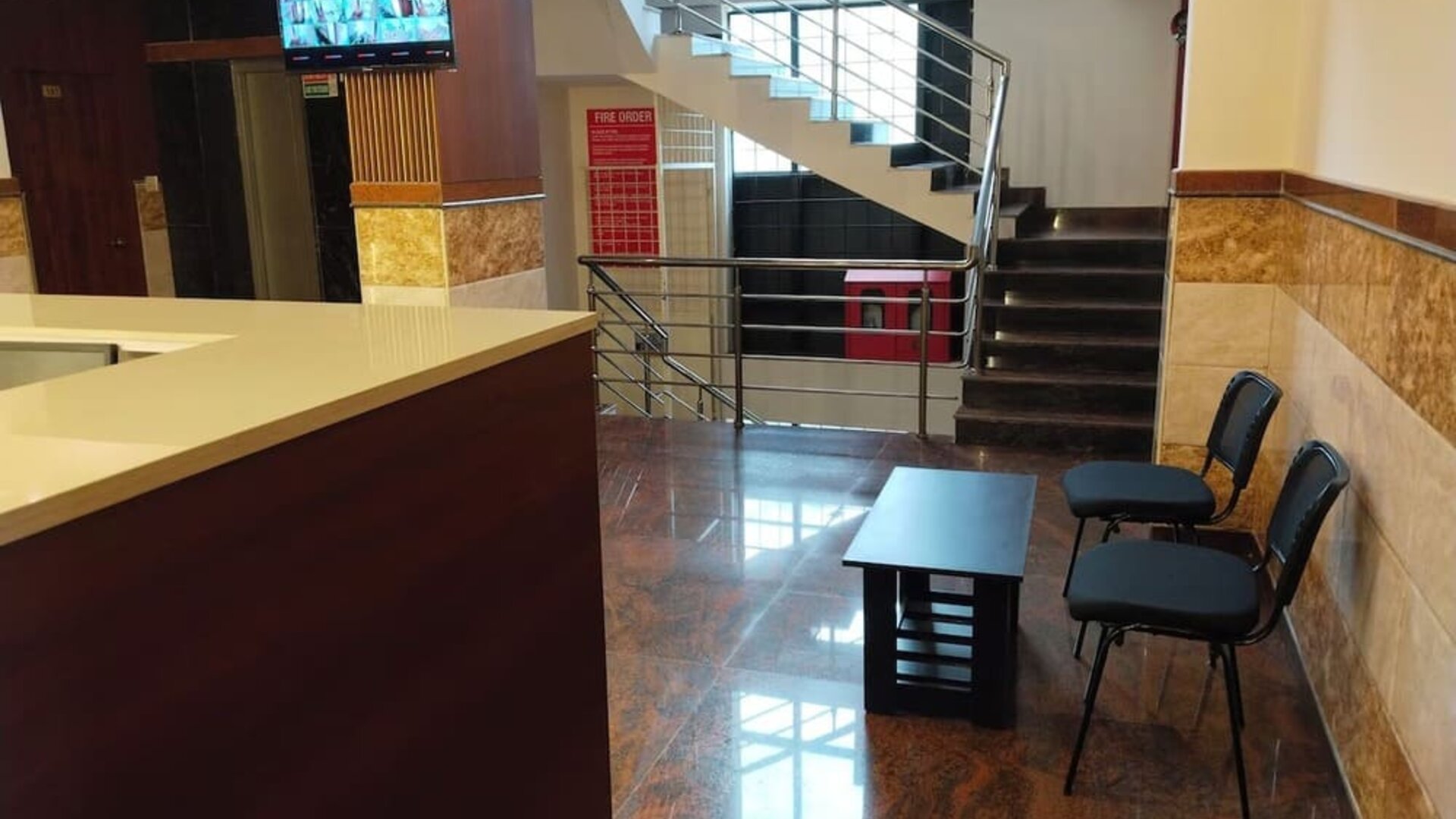 Welcoming hotel lobby featuring a polished reception counter, black guest chairs, coffee table, staircase with stainless steel railings, and glossy tiled flooring.