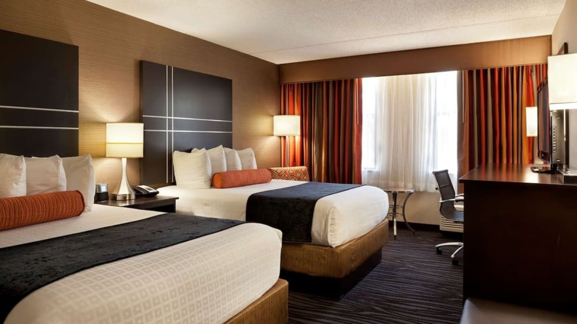 Guest room with two beds, crisp white bedding, dark bed runners, orange accent cushions, striped curtains, bedside lamps, and a work desk at Best Western Plus BWI Airport Arundel Mills.