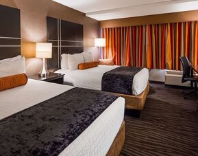 Guest room with two beds, white bedding, dark bed runners, orange accent cushions, striped curtains, bedside lamps, and a work desk at Best Western Plus BWI Airport Arundel Mills.
