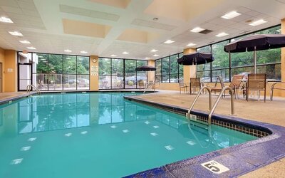 Indoor swimming pool with clear turquoise water, surrounding deck, metal handrails, large windows, and outdoor greenery visible through the glass at Best Western Plus BWI Airport Arundel Mills.