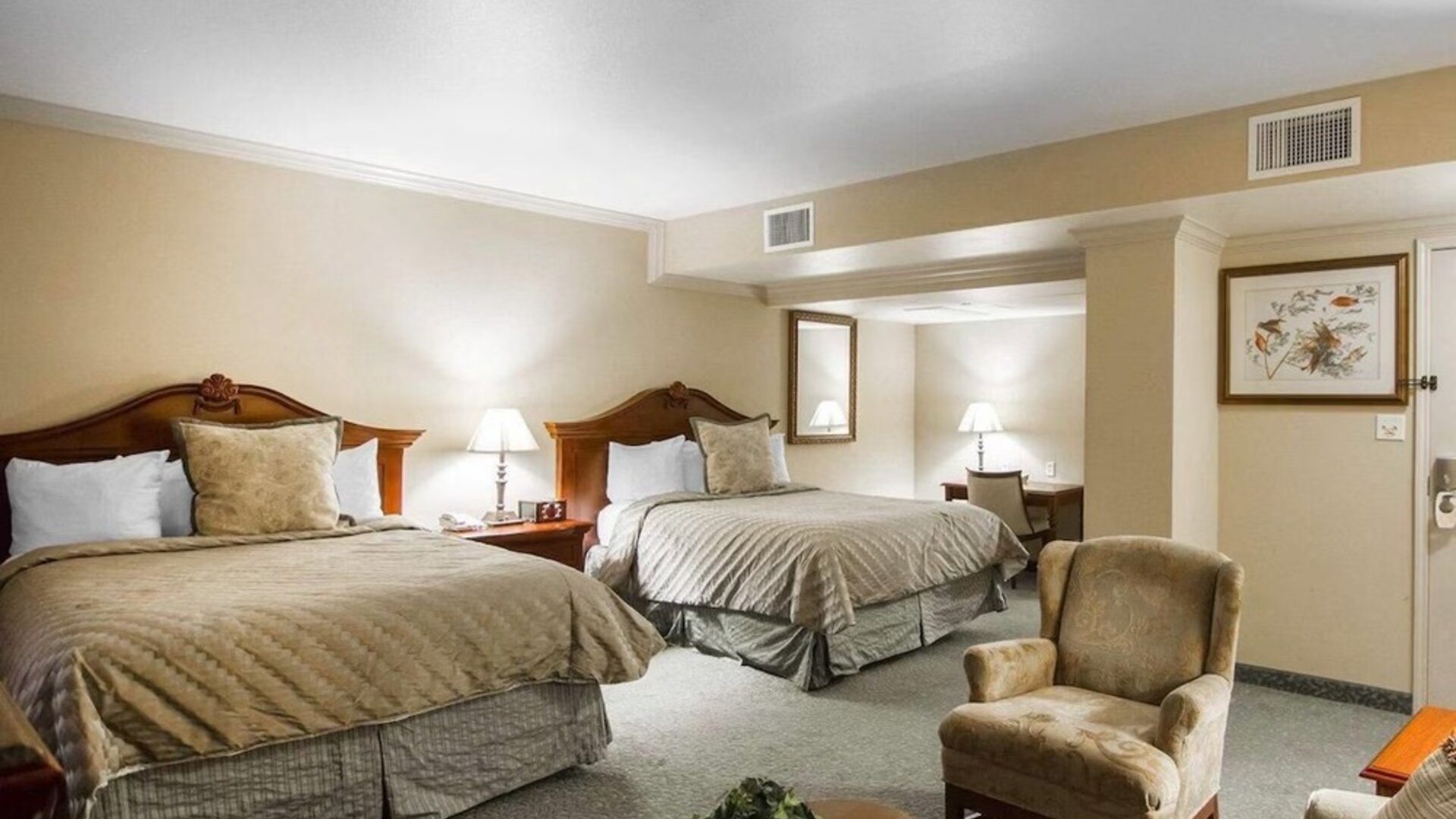 Spacious guest room featuring two beds with beige patterned bedding, wooden headboards, matching bedside tables, upholstered seating, and a carpeted floor at Liberty Hotel Cleburne, an Ascend Hotel Collection Member.