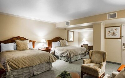 Spacious guest room featuring two beds with beige patterned bedding, wooden headboards, matching bedside tables, upholstered seating, and a carpeted floor at Liberty Hotel Cleburne, an Ascend Hotel Collection Member.