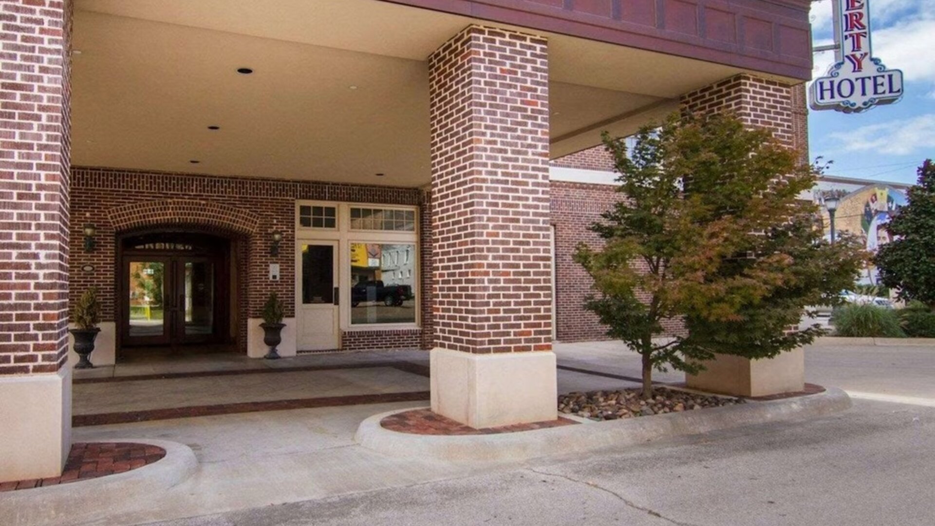 Exterior entrance of Liberty Hotel Cleburne, featuring a red-brick facade, covered porte-cochère, brick columns, landscaped planters, and the hotel's vertical sign.