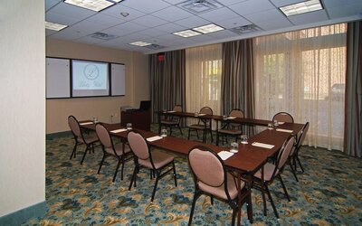 Meeting room arranged with conference tables and chairs in a U-shaped layout, a projection screen, presentation equipment, large curtained windows, and patterned carpeting at Liberty Hotel Cleburne, an Ascend Hotel Collection Member.