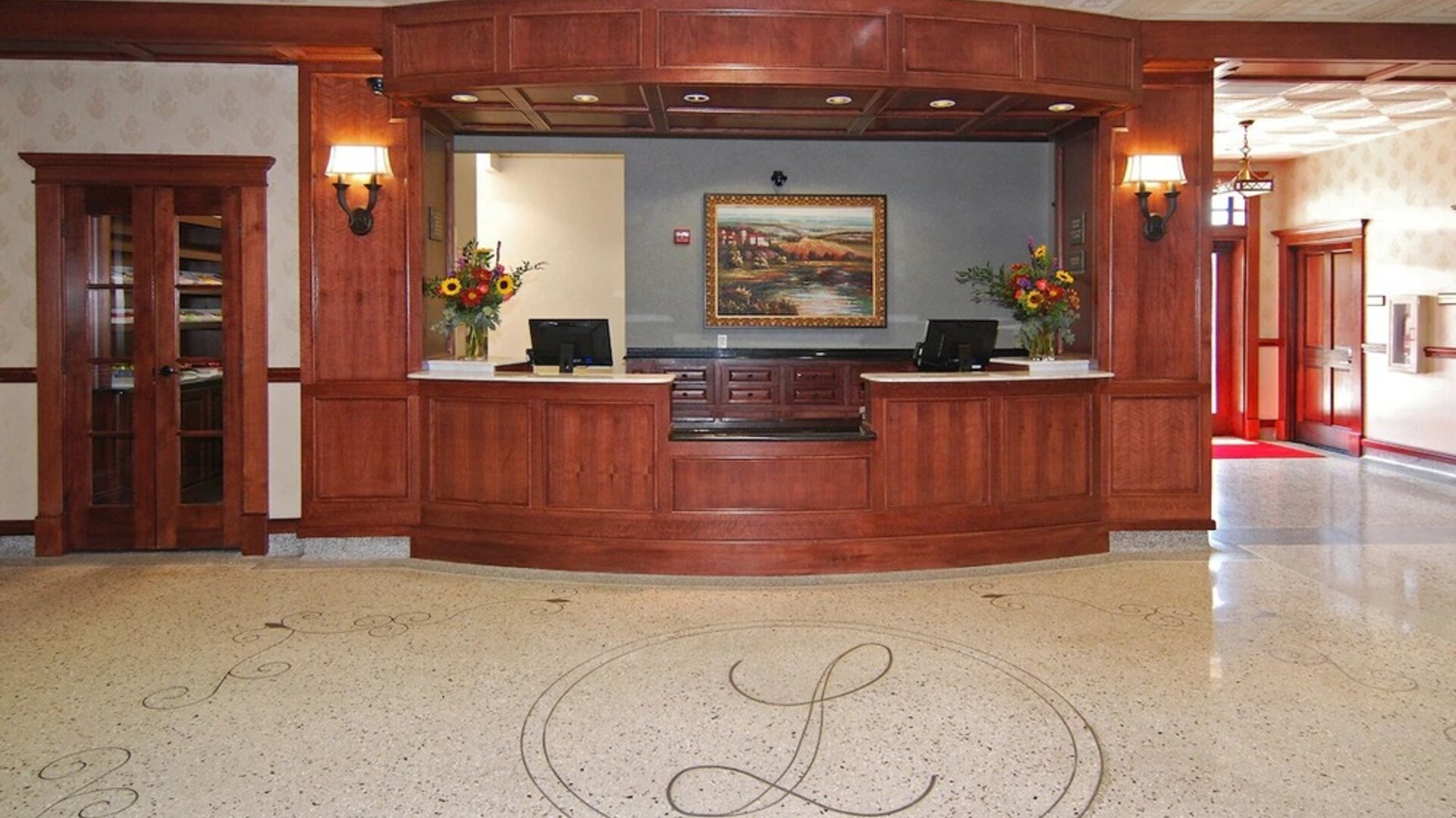 Hotel reception desk featuring rich wood paneling, two computer workstations, floral arrangements, decorative lighting, and a polished floor with an ornamental hotel emblem at Liberty Hotel Cleburne, an Ascend Hotel Collection Member.