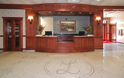 Hotel reception desk featuring rich wood paneling, two computer workstations, floral arrangements, decorative lighting, and a polished floor with an ornamental hotel emblem at Liberty Hotel Cleburne, an Ascend Hotel Collection Member.