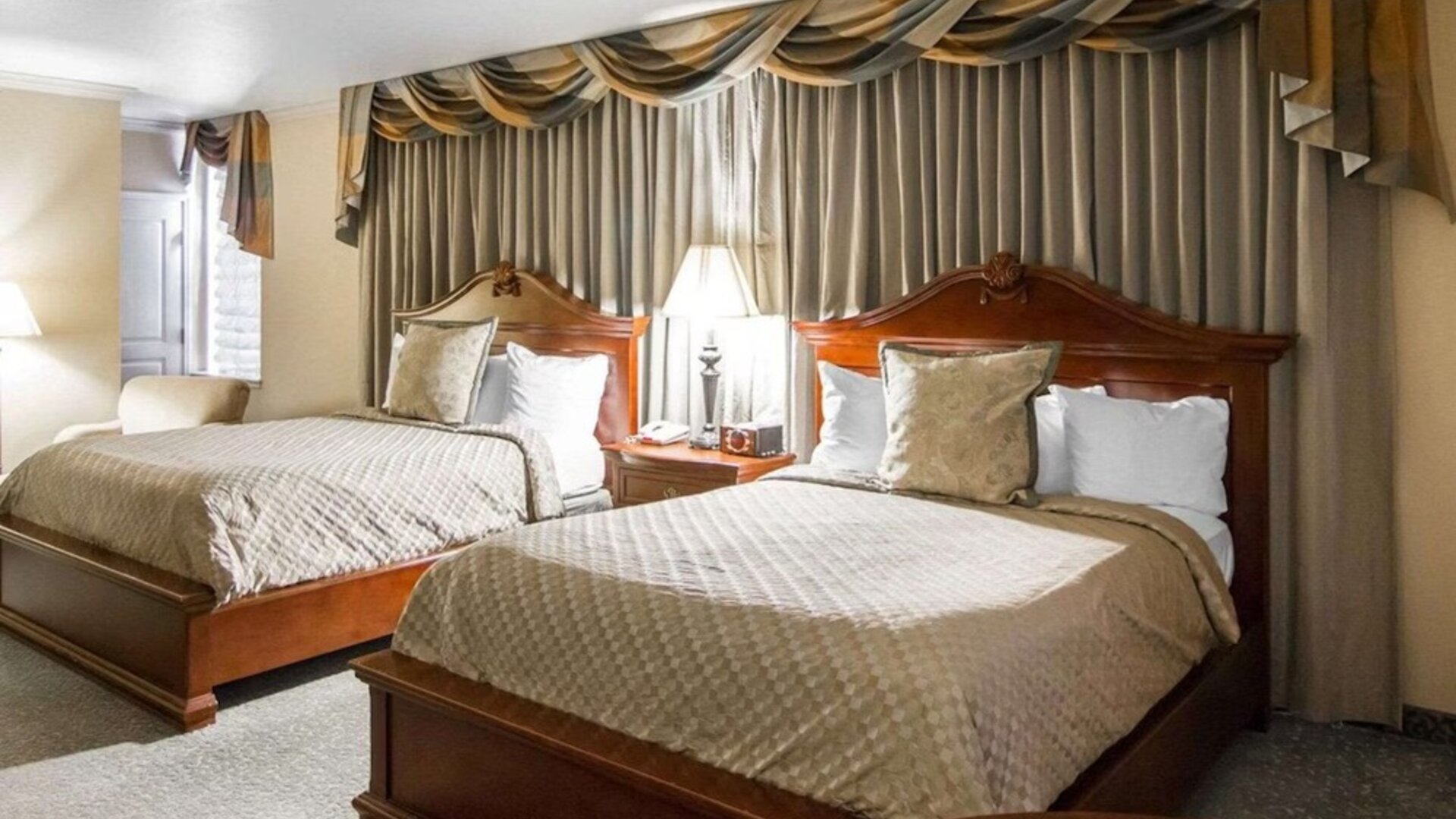 Guest room with two beds, beige patterned bedding, wooden headboards, matching bedside tables, decorative curtains, and a comfortable seating area at Liberty Hotel Cleburne, an Ascend Hotel Collection Member.