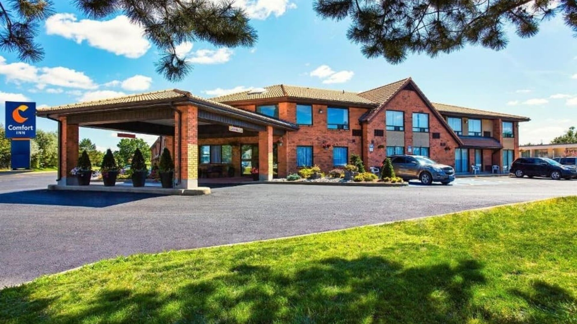 Exterior view of Comfort Inn Alma, showcasing its red-brick facade, covered entrance, blue hotel signage, parking area, and landscaped lawn.