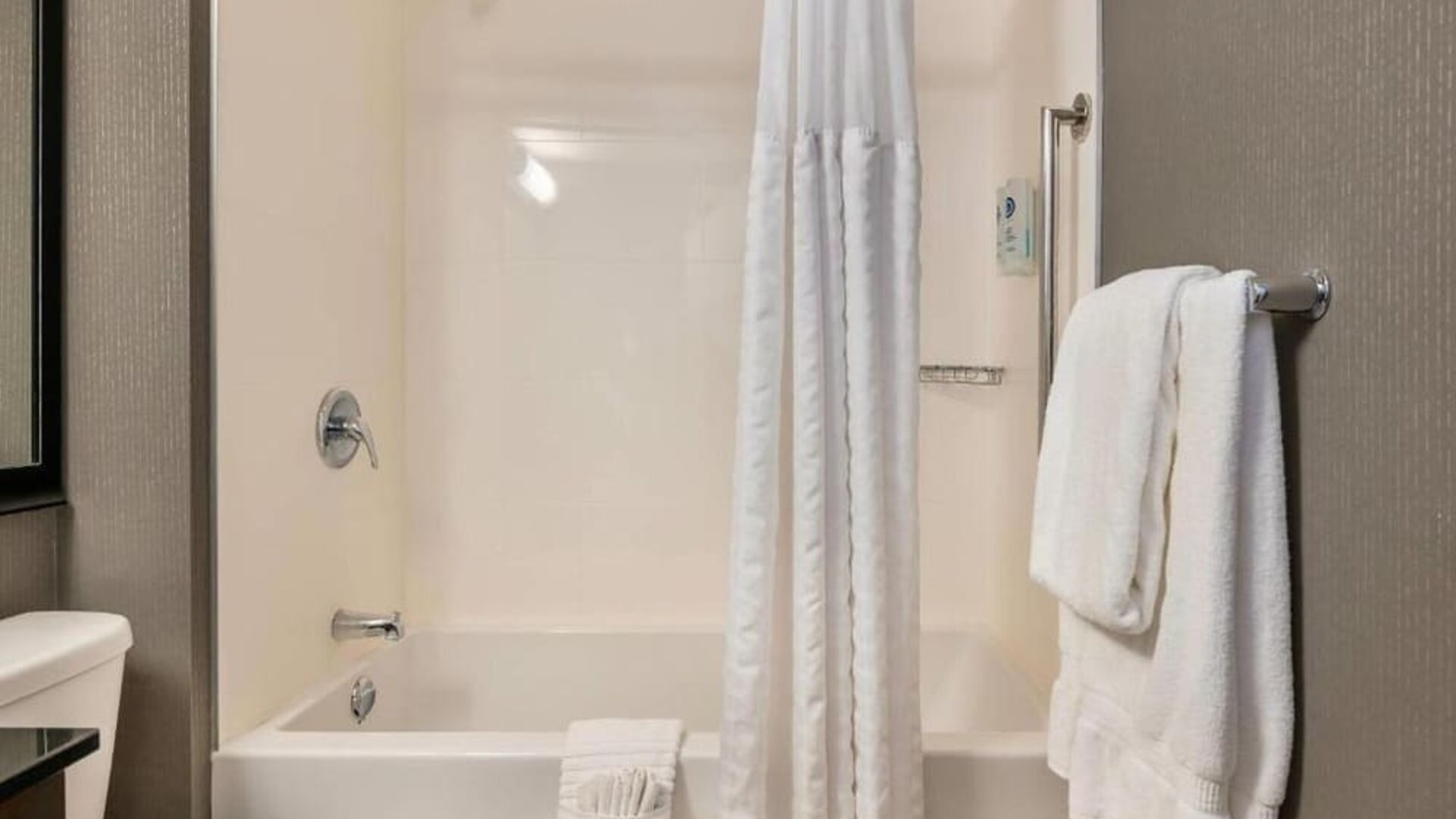 Guest bathroom with a white bathtub and shower curtain, chrome fixtures, wall-mounted grab bar, and neatly folded white towels at Comfort Inn Alma.