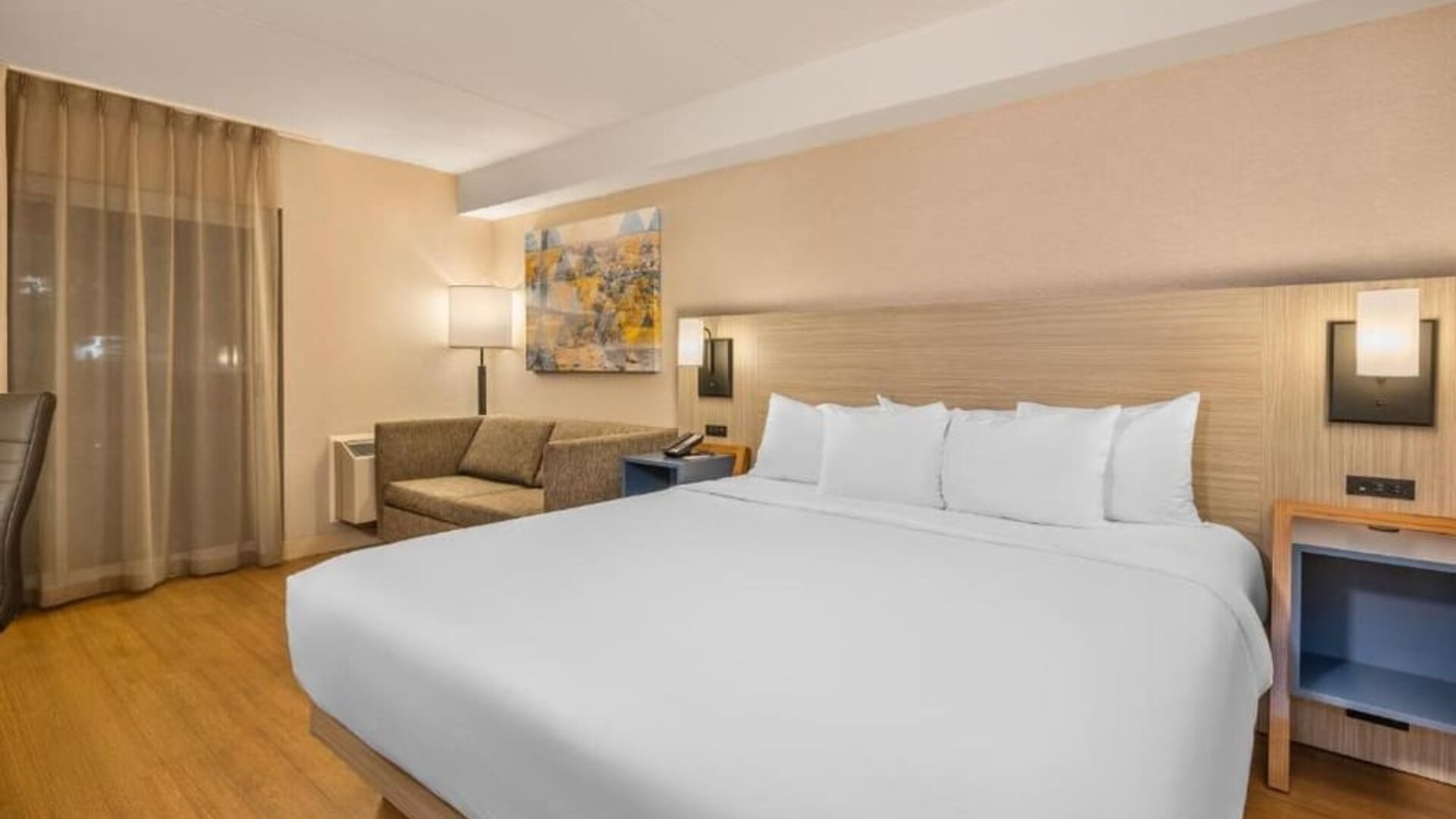 Contemporary king guest room featuring a large bed with white linens, a light wood headboard, a gray sofa, a desk and office chair, bedside shelving, and warm wood flooring at Comfort Inn Alma.