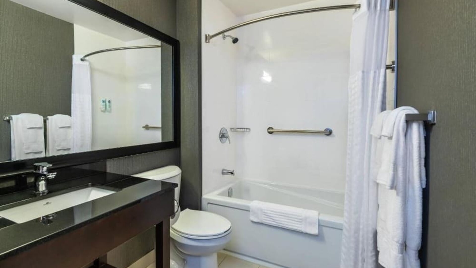Modern hotel bathroom featuring a white bathtub and shower curtain, granite vanity countertop, large black-framed mirror, toilet, chrome fixtures, and white towels at Comfort Inn Alma.