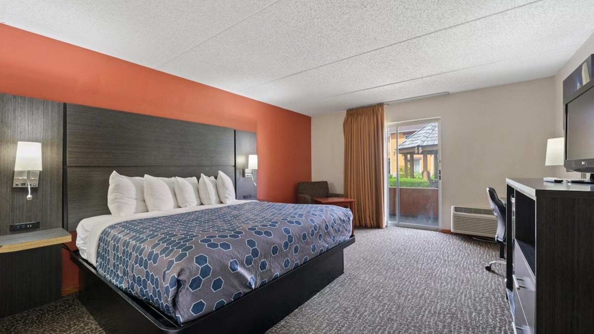Spacious king guest room featuring a king-size bed, blue geometric-patterned bedspread, dark wood furnishings, a desk and office chair, and sliding glass doors leading to a balcony at Chicago Club Inn & Suites.
