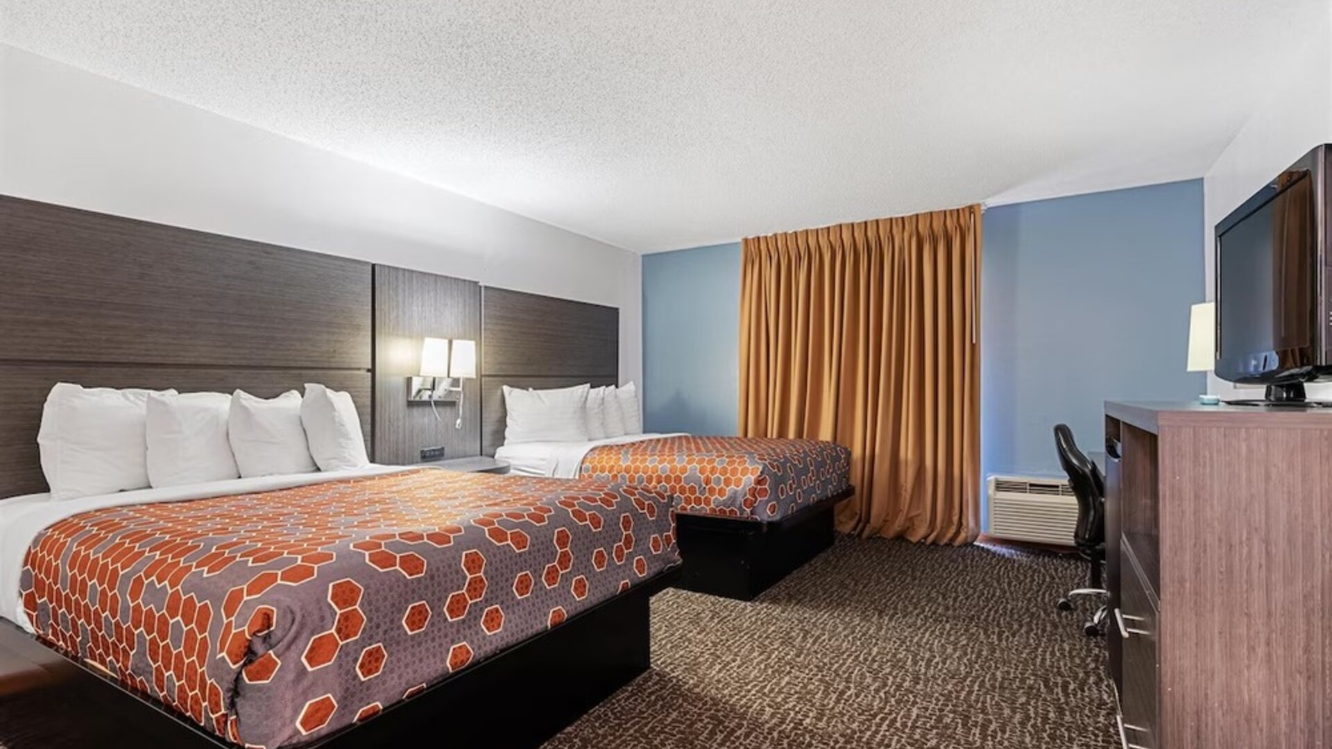 Guest room featuring two beds with orange geometric-patterned bedspreads, white pillows, a dark wood headboard, blue accent walls, and contemporary furnishings at Chicago Club Inn & Suites.