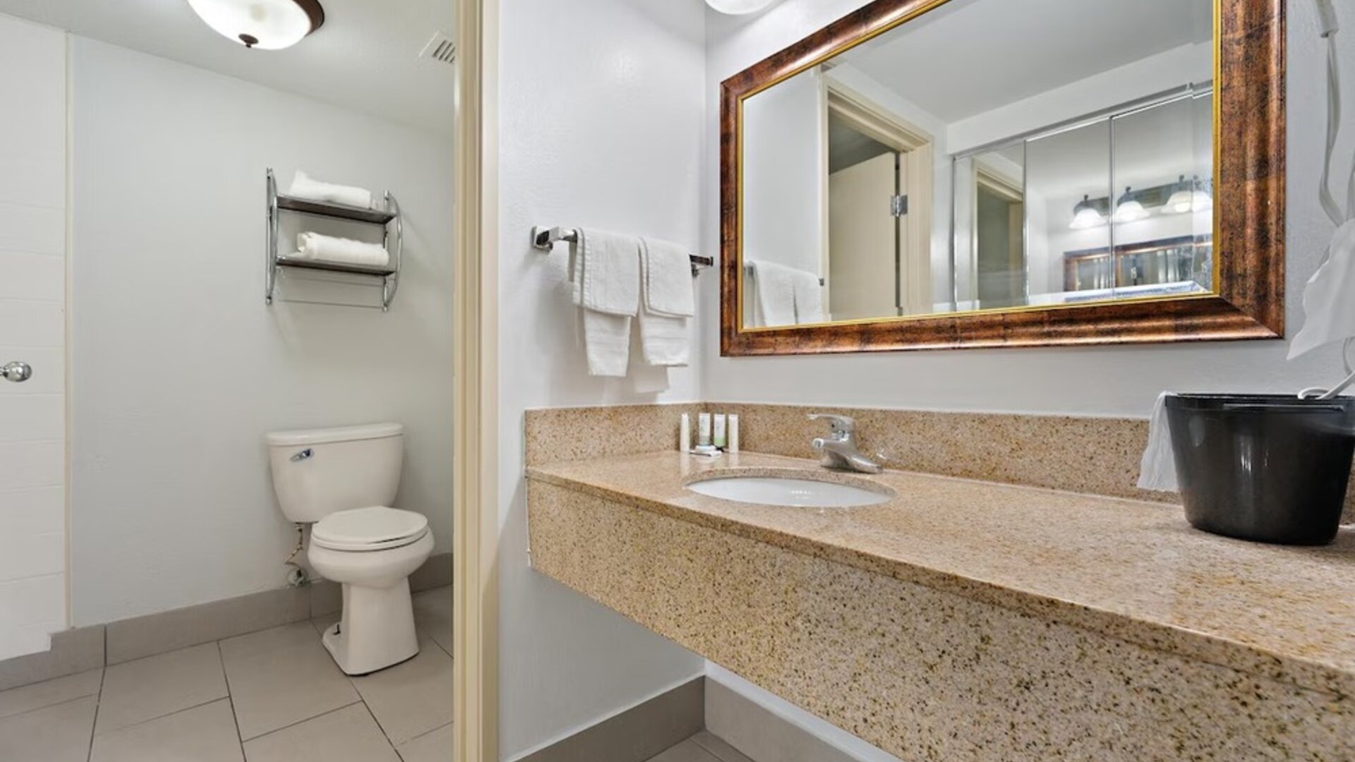 Guest bathroom featuring a beige granite vanity countertop, an inset sink, a large wood-framed mirror, white towels, a toilet, and tiled flooring at Chicago Club Inn & Suites.