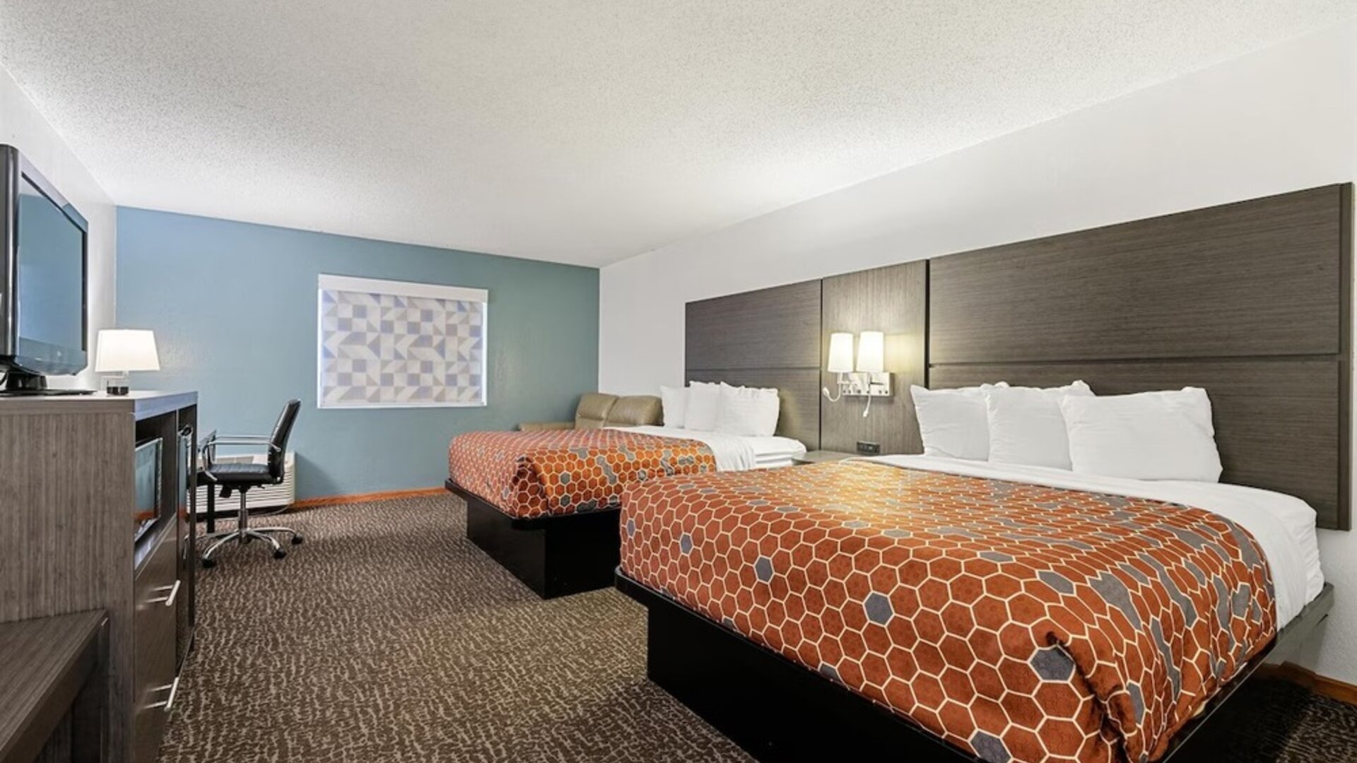 Guest room featuring two beds with orange geometric-patterned bedspreads, white pillows, a dark wood headboard, a television, a desk, and a seating area at Chicago Club Inn & Suites.