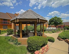 Exterior courtyard featuring a wooden gazebo, landscaped gardens, paved walkways, outdoor seating, and the hotel's orange-colored buildings at Chicago Club Inn & Suites.