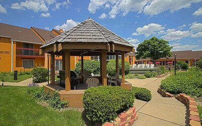 Exterior courtyard featuring a wooden gazebo, landscaped gardens, paved walkways, outdoor seating, and the hotel's orange-colored buildings at Chicago Club Inn & Suites.