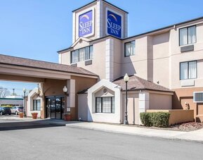 Daytime exterior view of the Sleep Inn hotel featuring a multi-story beige building, prominent blue Sleep Inn signage, covered entrance with glass doors, decorative exterior lighting, landscaped shrubs, and a paved driveway.