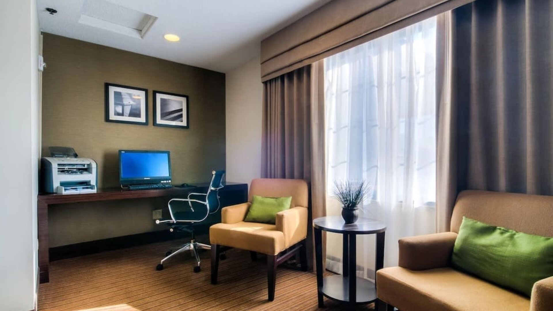 Cozy hotel business center featuring a desktop computer, printer, dark wooden workstation, ergonomic office chair, two beige upholstered armchairs with green accent pillows, a round side table, and a large window providing natural light.