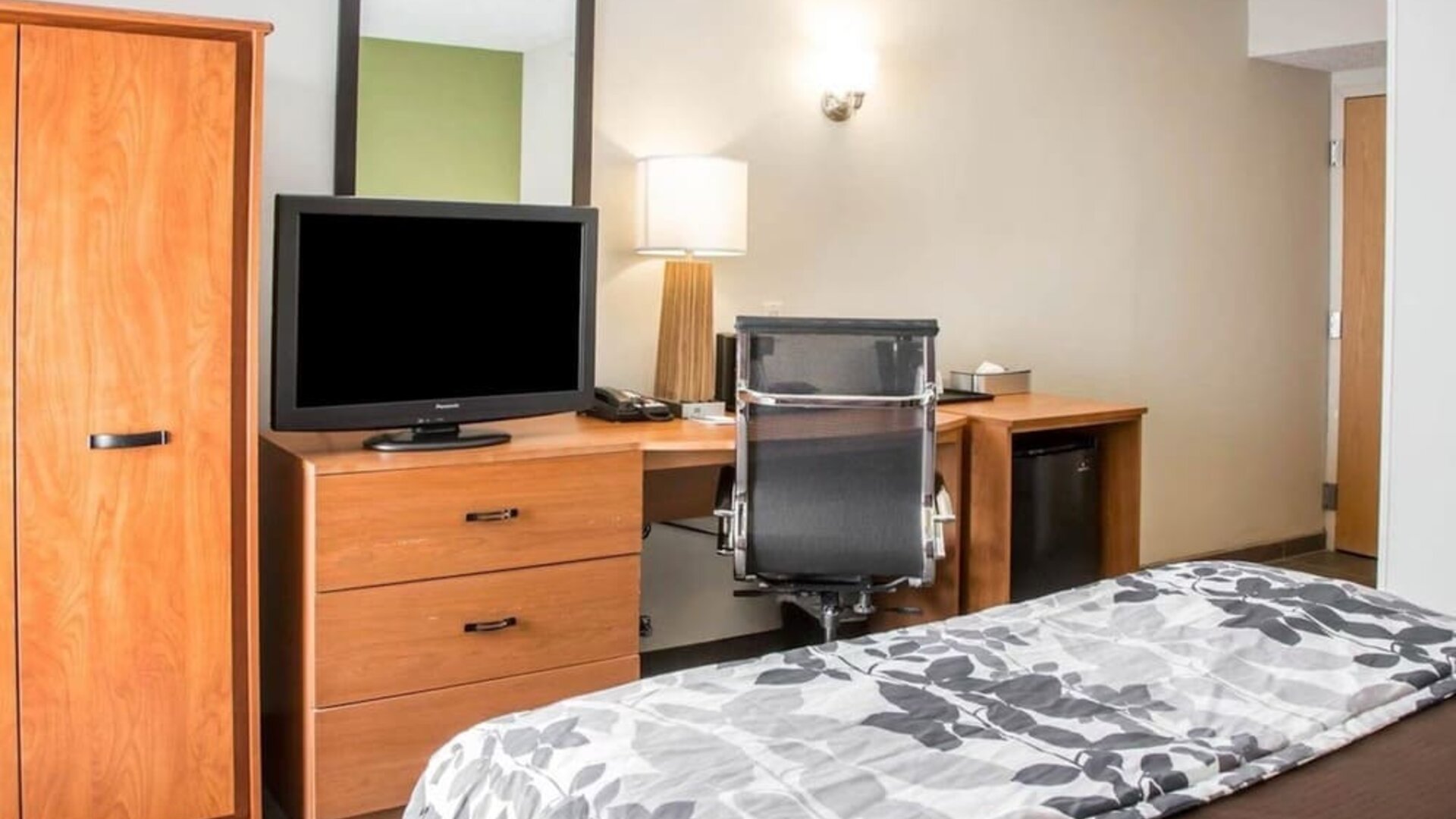 Sleep Inn guest room featuring a wooden dresser with flat-screen television, spacious work desk with black ergonomic office chair, large wall mirror, bedside lamp, wooden wardrobe, mini refrigerator, and a bed with brown bedding and a gray floral-patterne