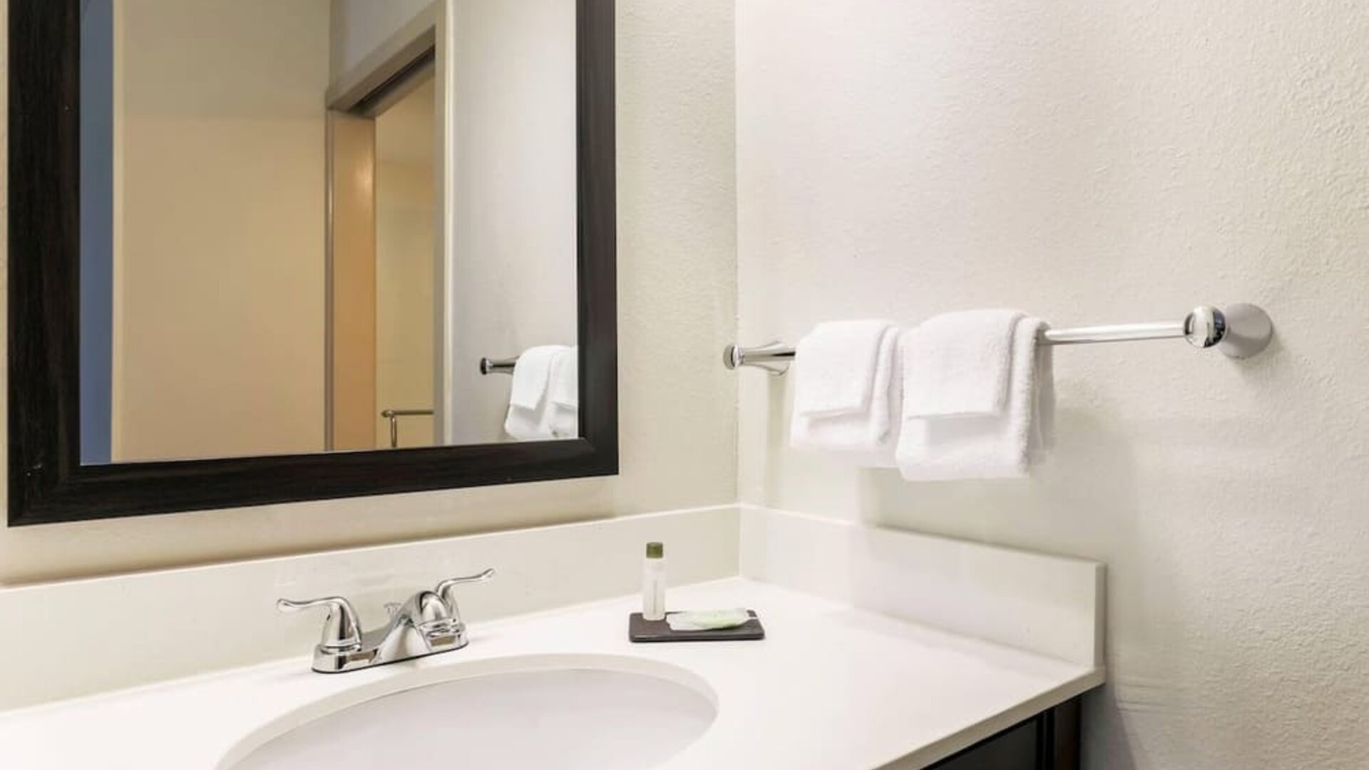Clean hotel bathroom vanity featuring a white countertop with an integrated sink, chrome faucet, large black-framed mirror, neatly folded white hand towels, wall-mounted towel rack, and complimentary toiletries.