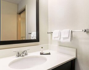 Clean hotel bathroom vanity featuring a white countertop with an integrated sink, chrome faucet, large black-framed mirror, neatly folded white hand towels, wall-mounted towel rack, and complimentary toiletries.
